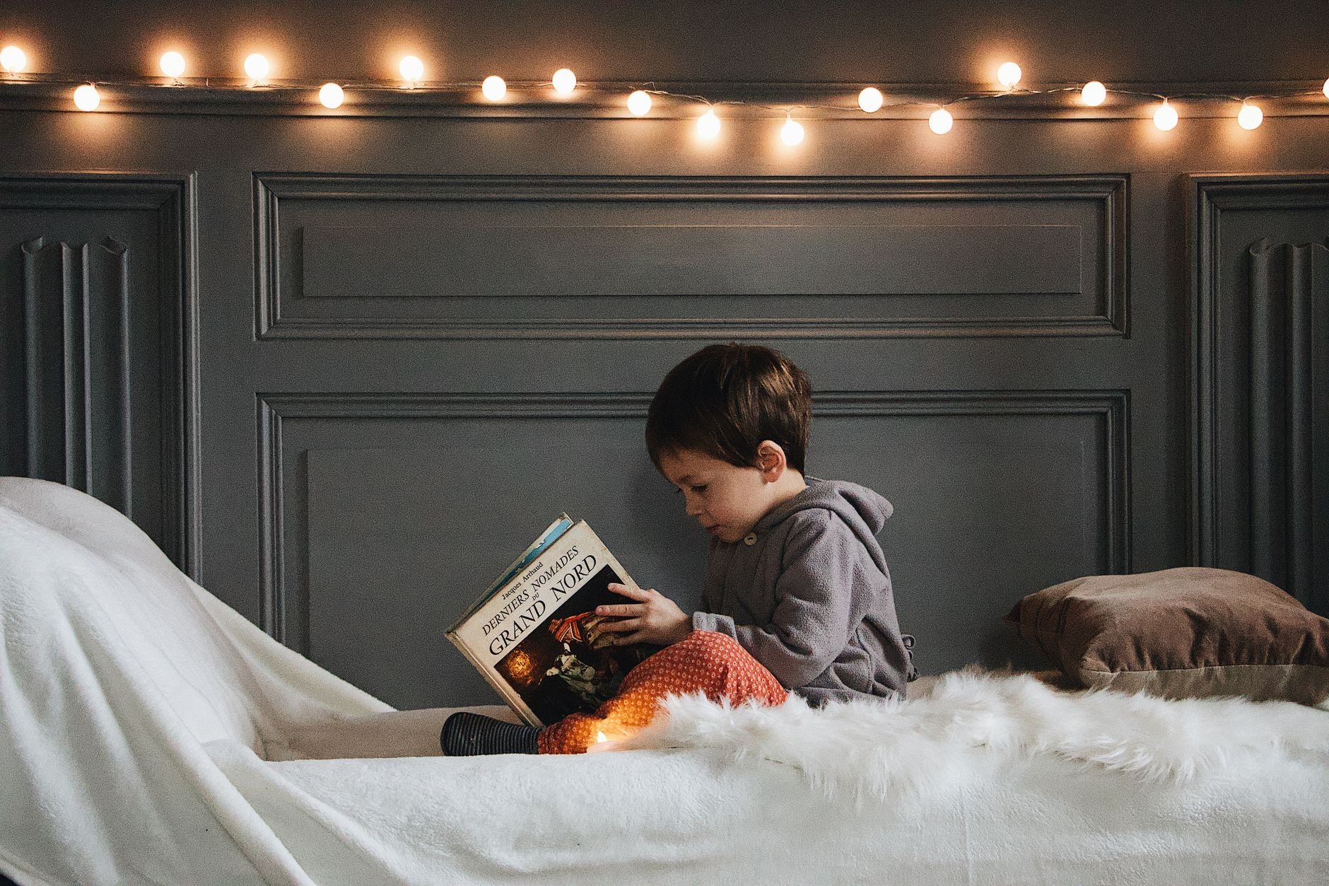 A young boy reading a book in a cozy bedroom with warm string lights and soft textures